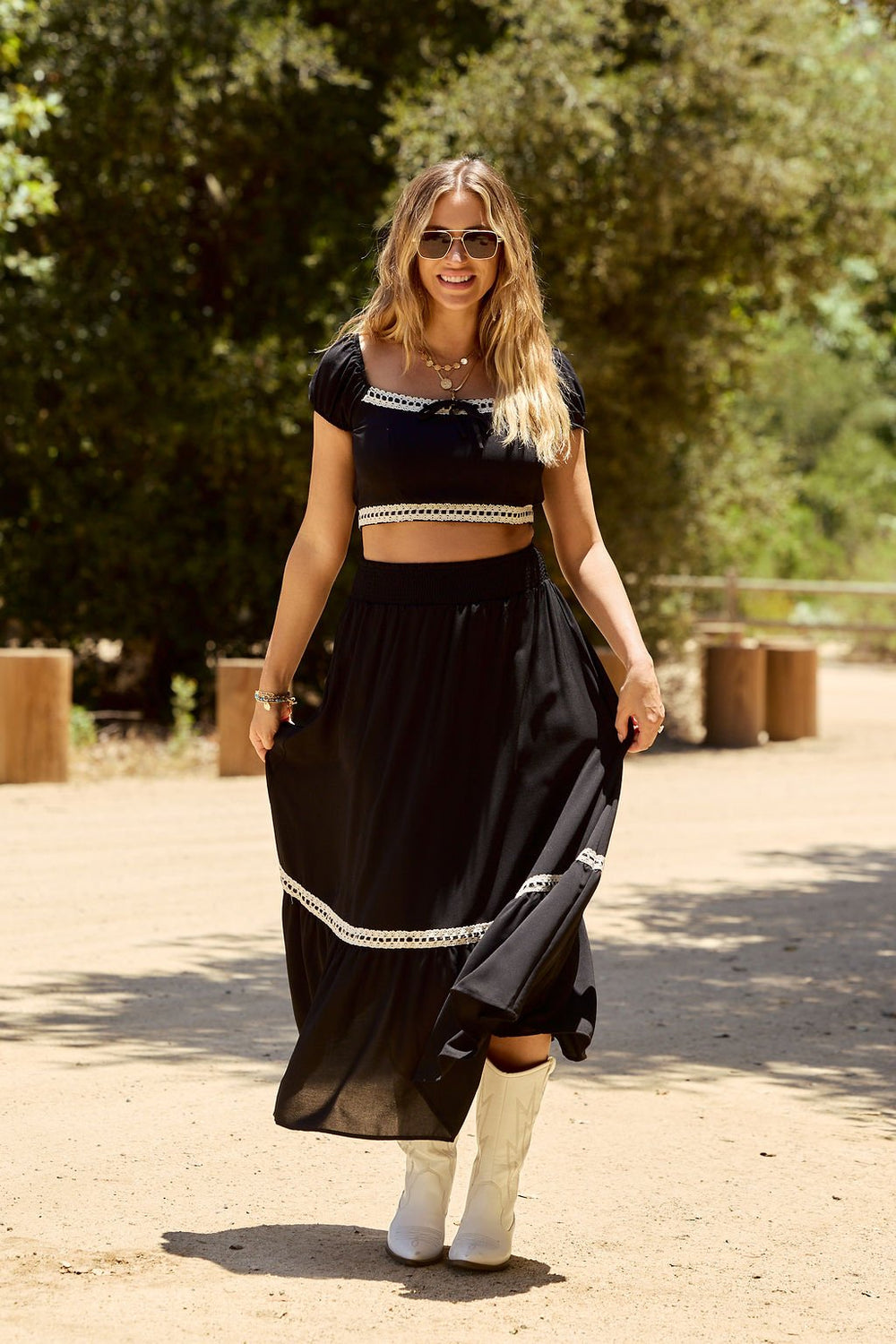 Woman outdoors in sunglasses, black crop top, white cowboy boots, smiling in the Jolene Black Lace Trim Maxi Skirt.