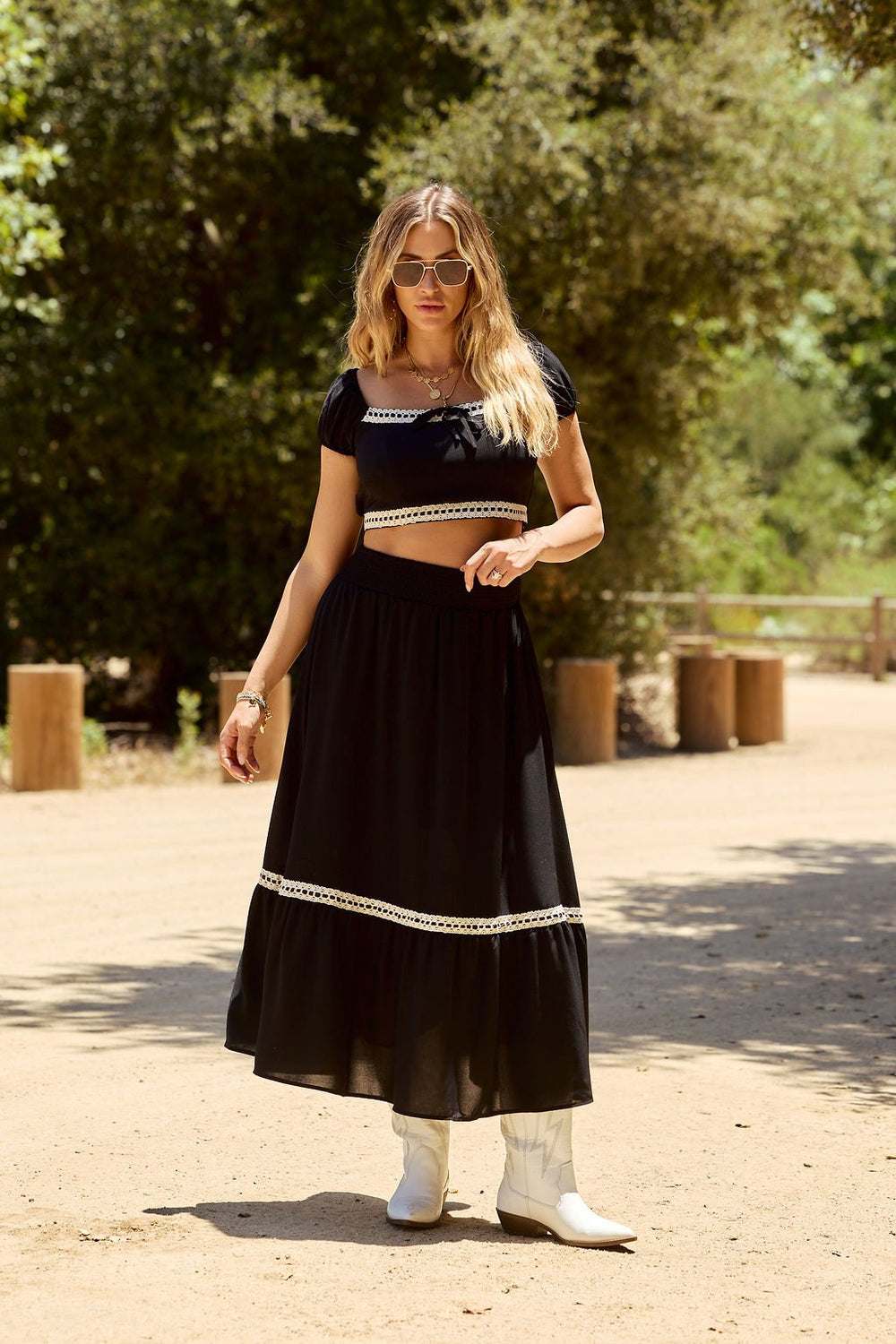 Woman outdoors in the Jolene Black Lace Trim Maxi Skirt, black crop top, white boots, and sunglasses on a dirt path.