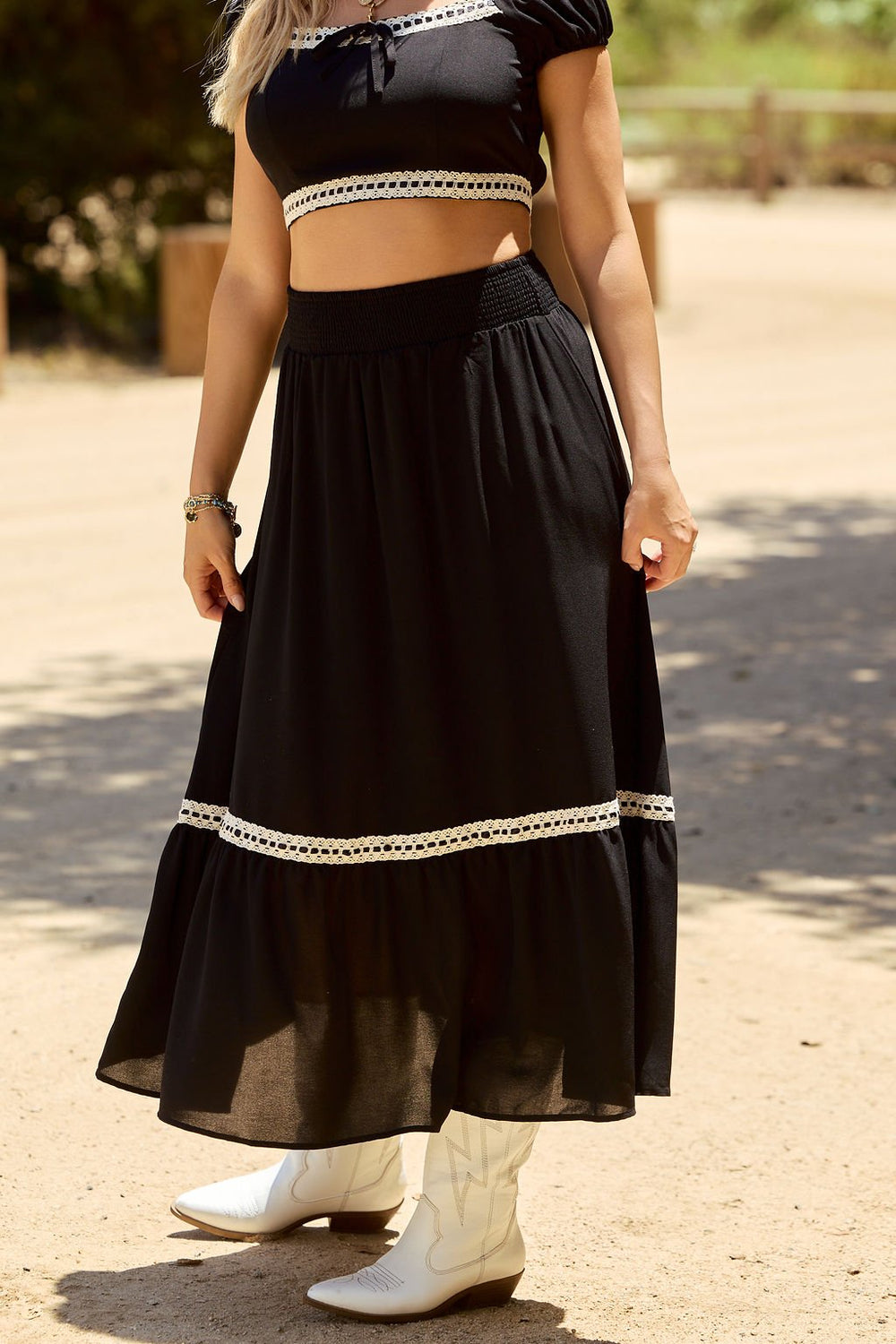 Woman wearing the Jolene Black Lace Trim Maxi Skirt, styled with a black crop top and white cowboy boots, standing outdoors.