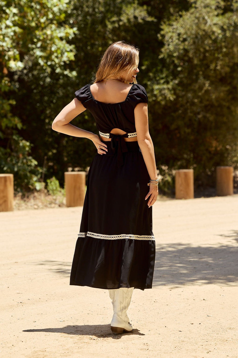 A woman wearing the Jolene Black Lace Trim Maxi Skirt and white boots stands outdoors on a sunny day, facing away from the camera.