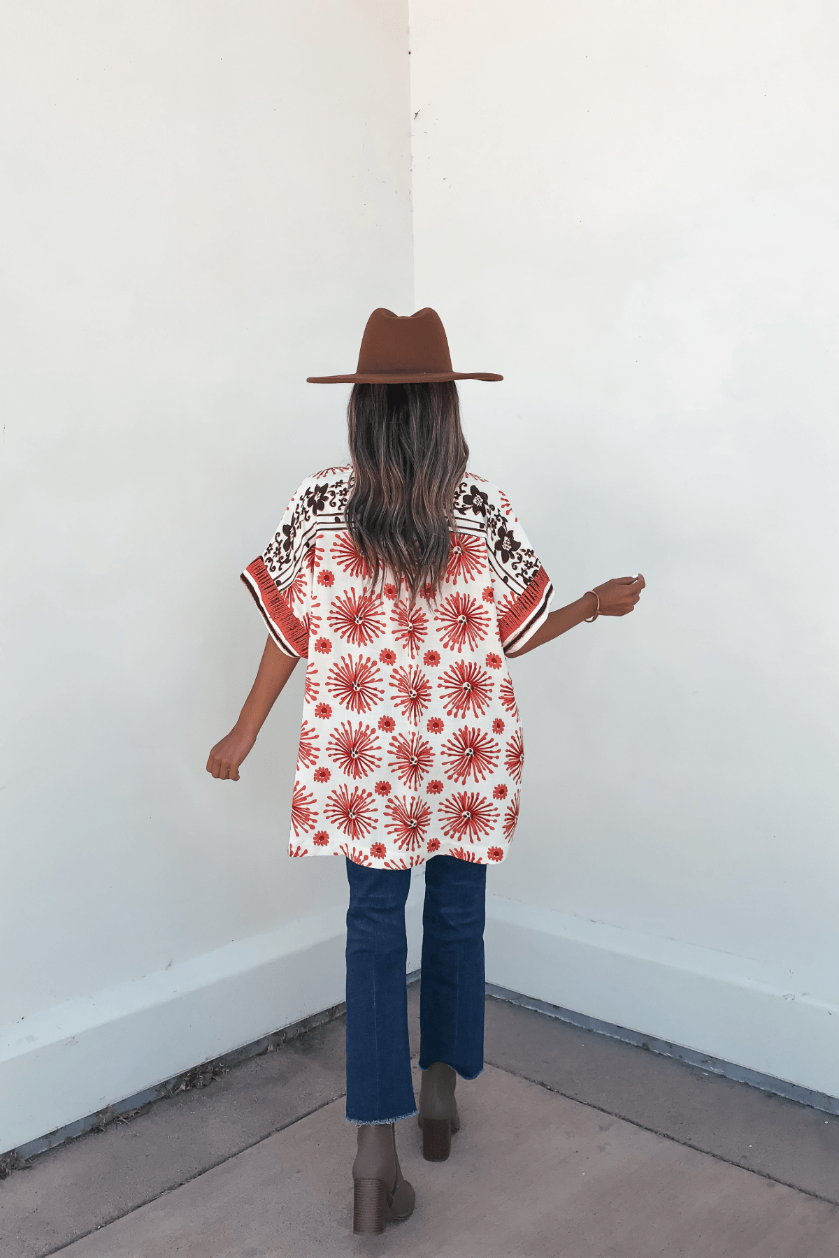 A woman in the Jolene Multi Red Border Print Kimono Top stands facing a white wall, styled with a brown hat and blue jeans.