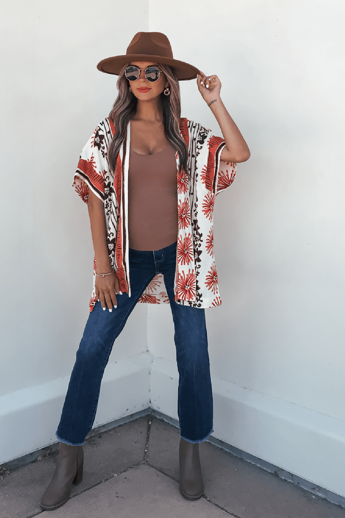 Woman wears Jolene Multi Red Border Print Kimono Top, brown hat, sunglasses, brown top, blue jeans, and boots by a white wall.