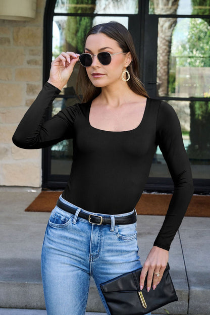 Woman in sunglasses wears the Jordan Black Square Neck Bodysuit with jeans and holds a black clutch outside a stone-walled building.