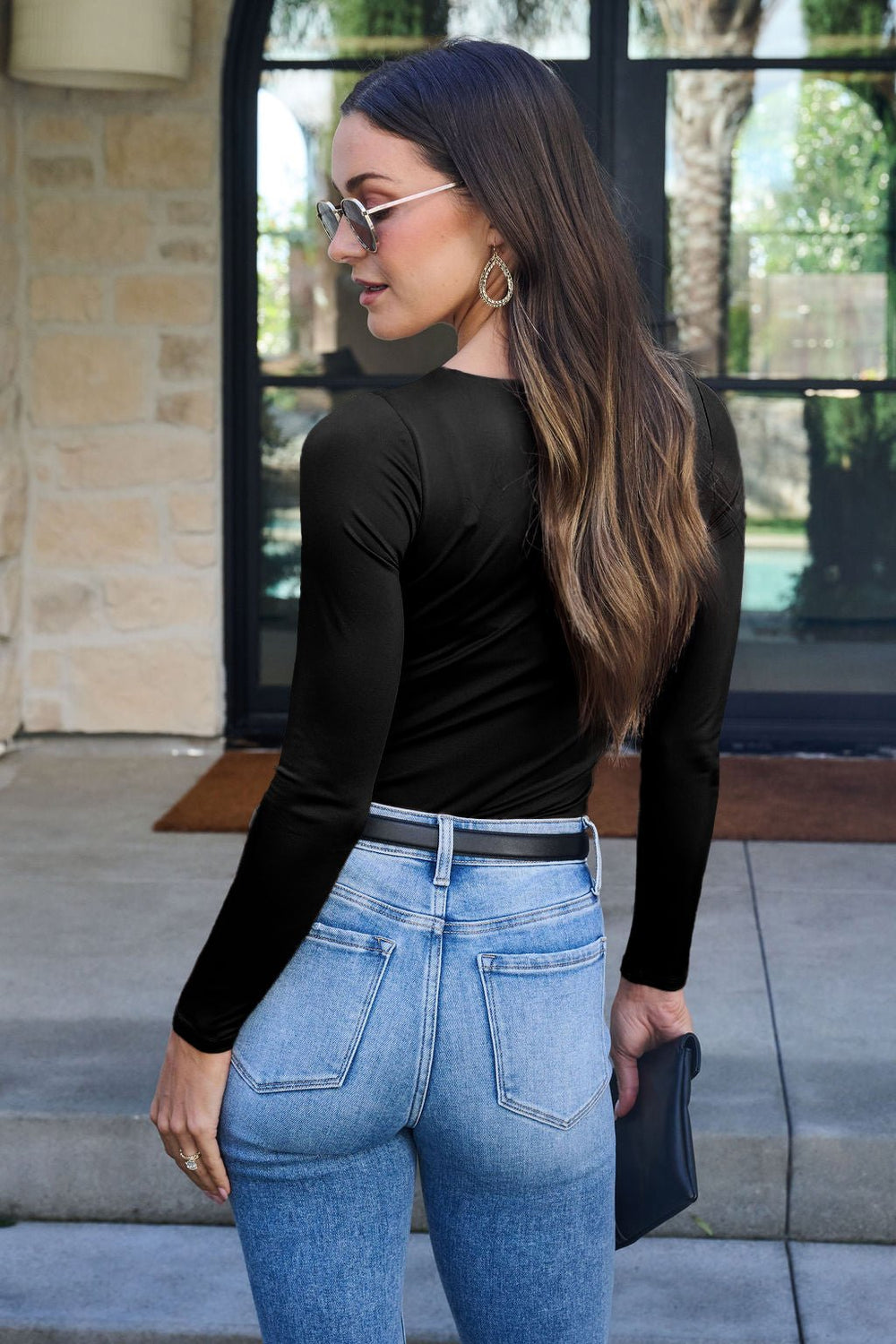 A woman in sunglasses and blue jeans models the Jordan Black Square Neck Bodysuit outdoors, facing away and looking over her shoulder.