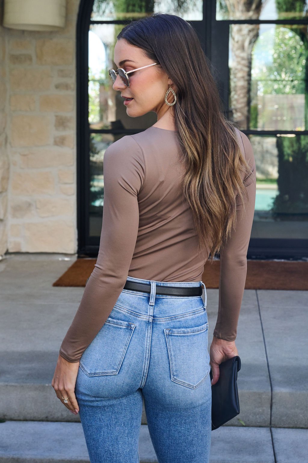 A woman in sunglasses and the Jordan Mocha Square Neck Bodysuit with blue jeans stands outdoors, facing away.