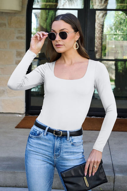 Woman in sunglasses and jeans wearing the Jordan White Square Neck Bodysuit, holding a black clutch outside glass doors.