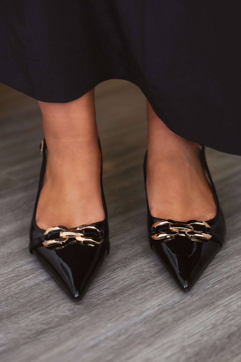 Someone wearing Josephine Black Slingback Kitten Heels - FINAL SALE, featuring gold chain accents, stands on a wooden floor.