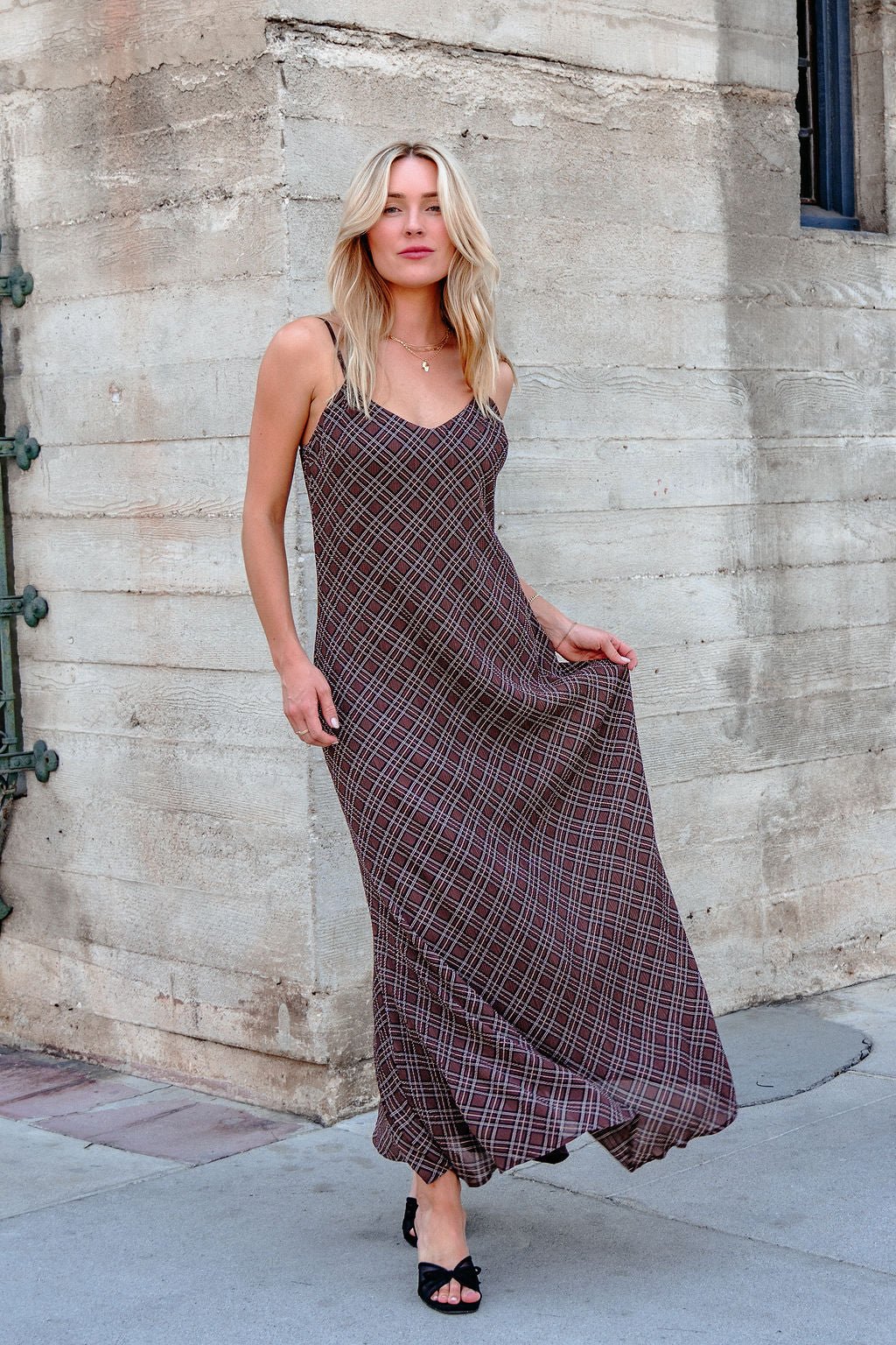 A woman wears the Josie Brown Plaid Maxi Dress - DOORBUSTER with black heels, standing by a beige stone wall on the sidewalk.