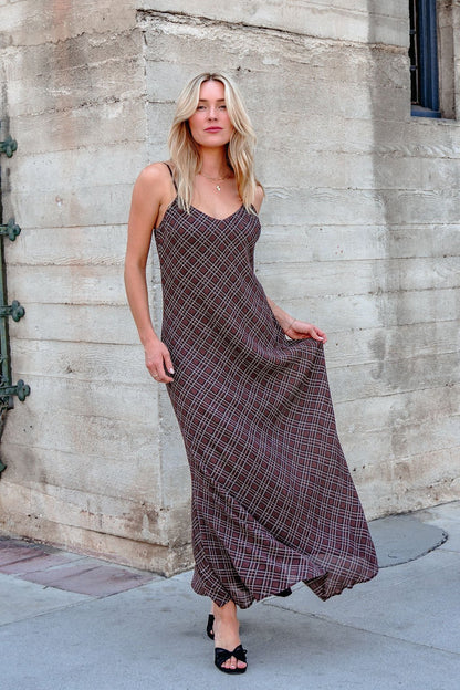A woman wears the Josie Brown Plaid Maxi Dress - DOORBUSTER with black heels, standing by a beige stone wall on the sidewalk.