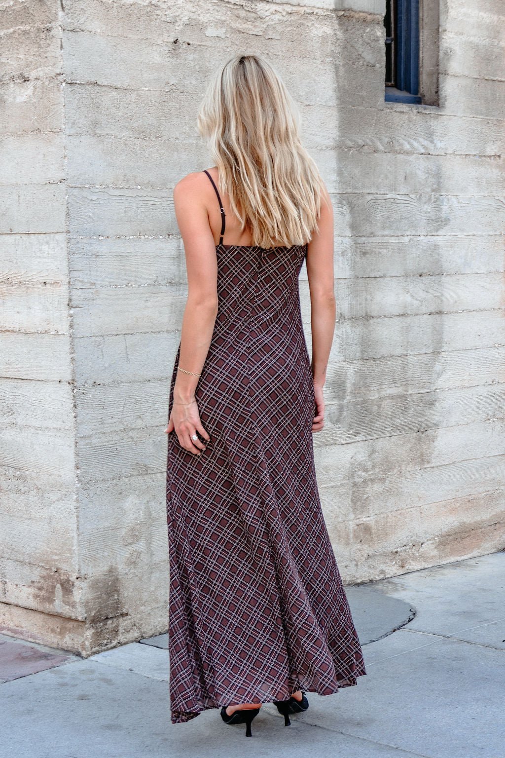 A woman with long blonde hair wears the Josie Brown Plaid Maxi Dress - DOORBUSTER, facing a textured concrete wall.