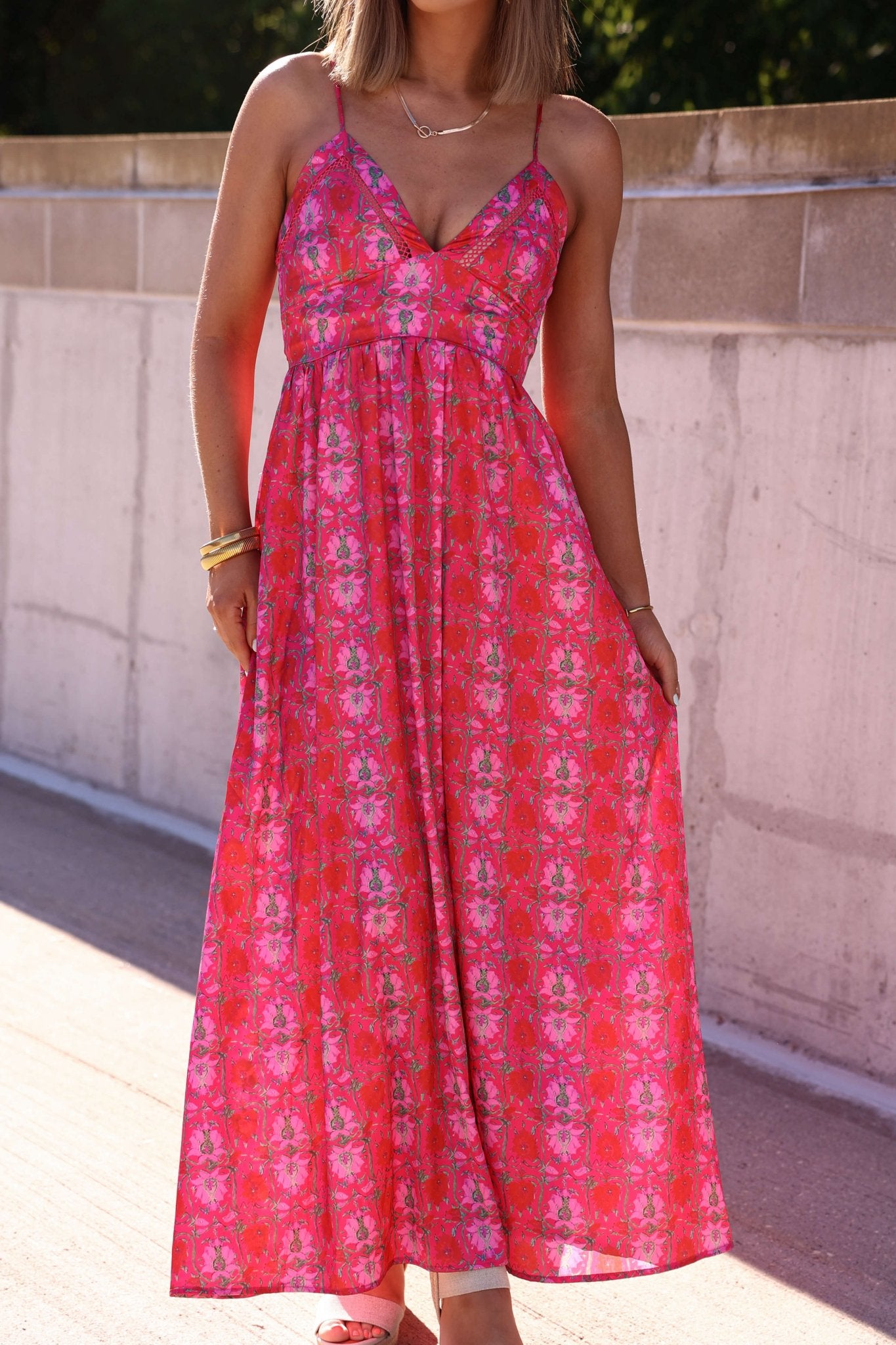 A woman in the Joyful Pink Floral Satin Maxi Dress stands outdoors by a concrete wall.