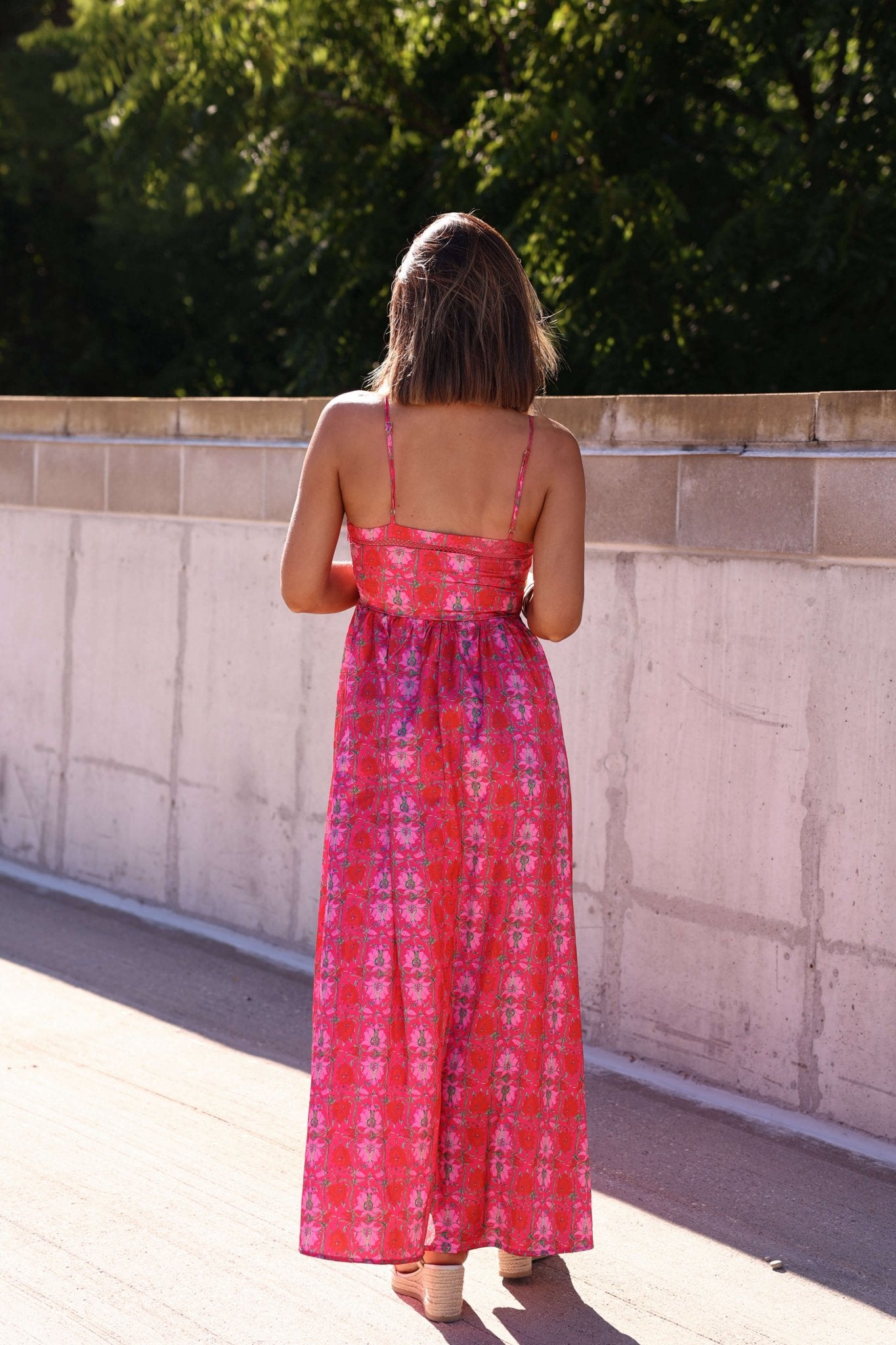 A woman in the Joyful Pink Floral Satin Maxi Dress stands outdoors with her back to the camera, facing greenery and a concrete wall.