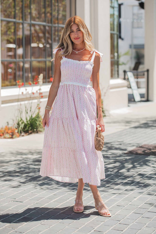 Woman in a Juliette Crochet Tiered Midi Dress - Pink and sandals stands on a sunny sidewalk, holding a woven handbag.