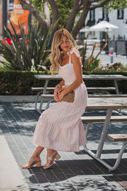 A woman in a Juliette Crochet Tiered Midi Dress - Pink sits outdoors on a picnic table, holding a woven clutch and smiling.