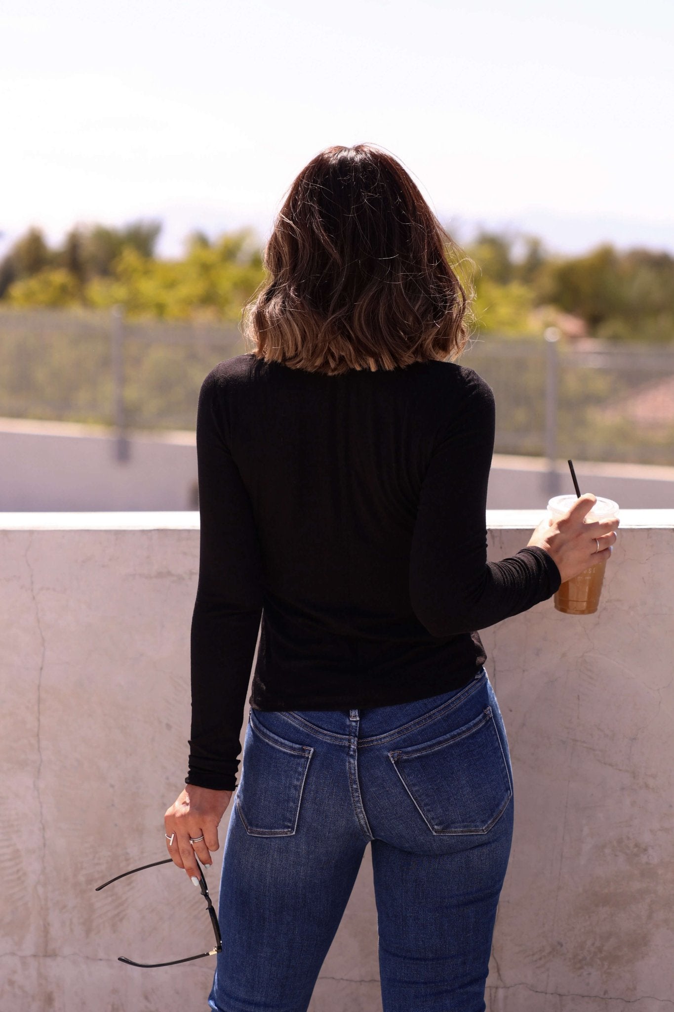 A woman with wavy hair outdoors in fall holds iced coffee and sunglasses, wearing the Juniper Black Ribbed Crewneck Top.