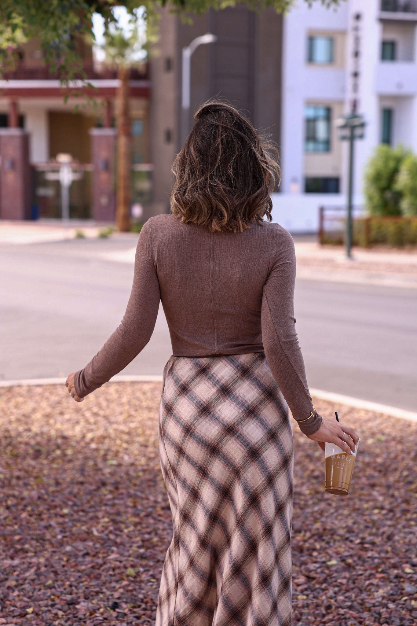 A woman with wavy hair walks outside, holding iced coffee and wearing the Juniper Hazelnut Ribbed Crewneck Top—perfect fall style.