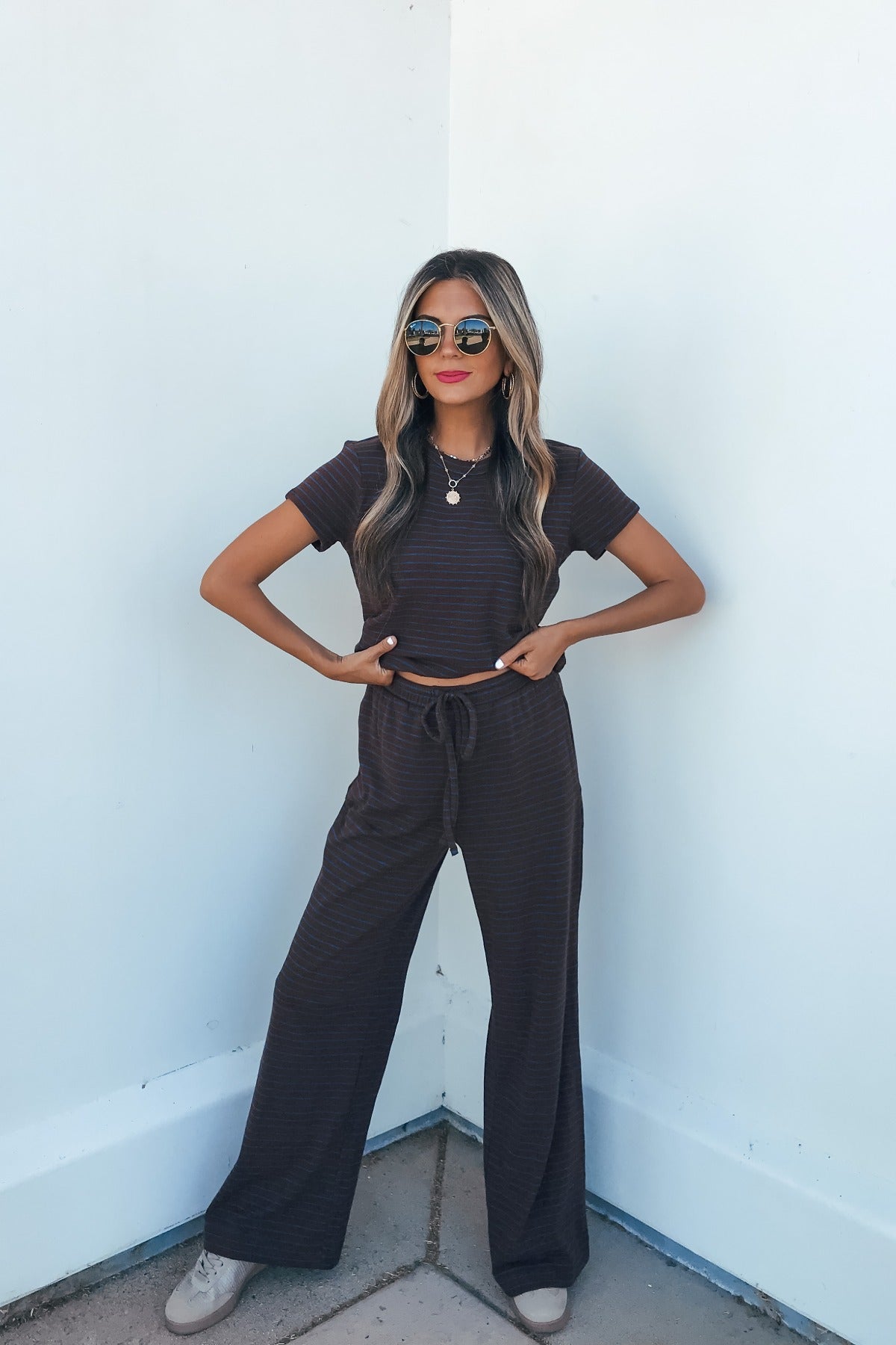 Woman wears Kacey Striped Wide Leg Pants in brown, sunglasses, and a matching set, standing confidently against a white wall.