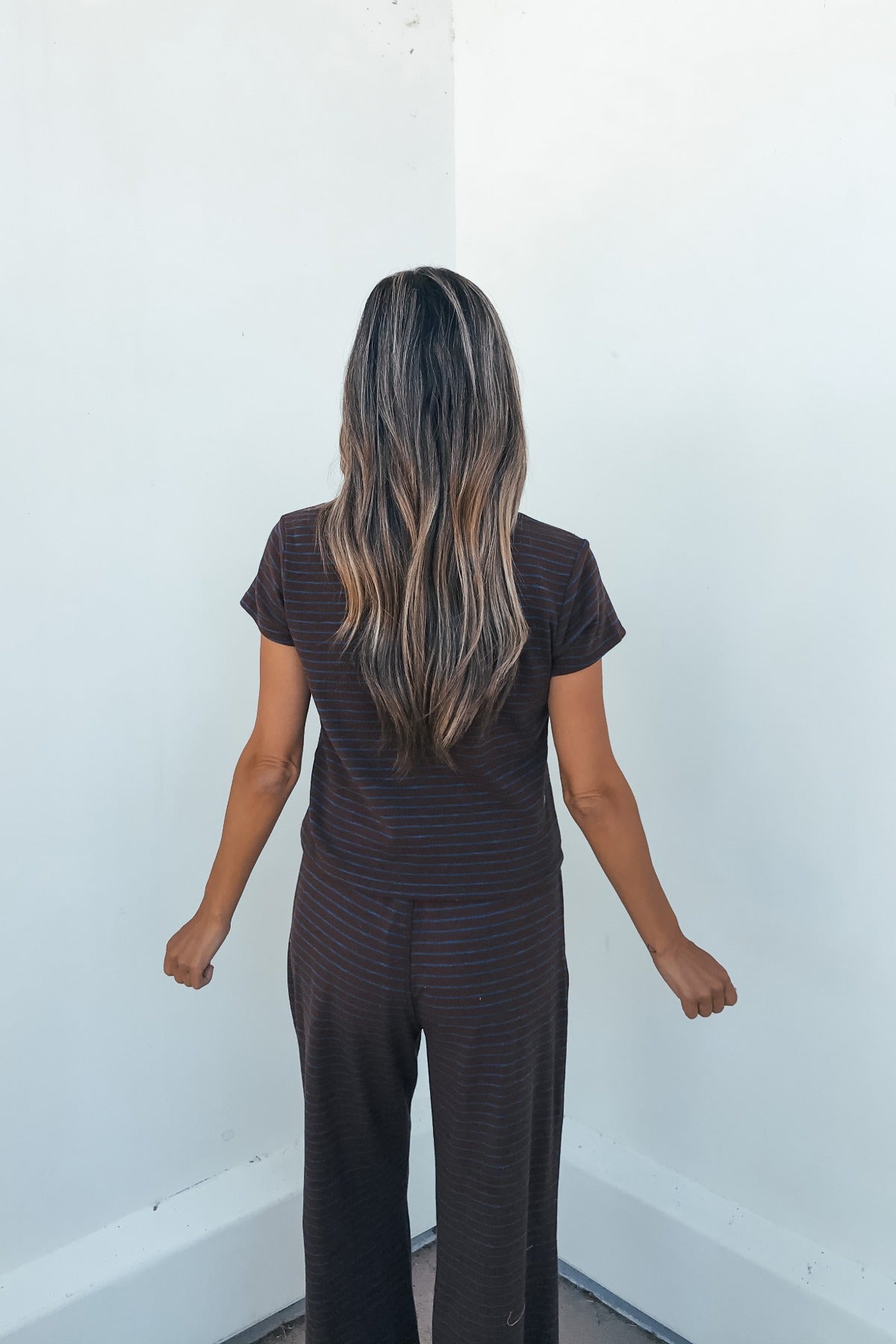 Woman with long, wavy brown hair wears Kacey Striped Wide Leg Pants - Brown, facing a white wall.