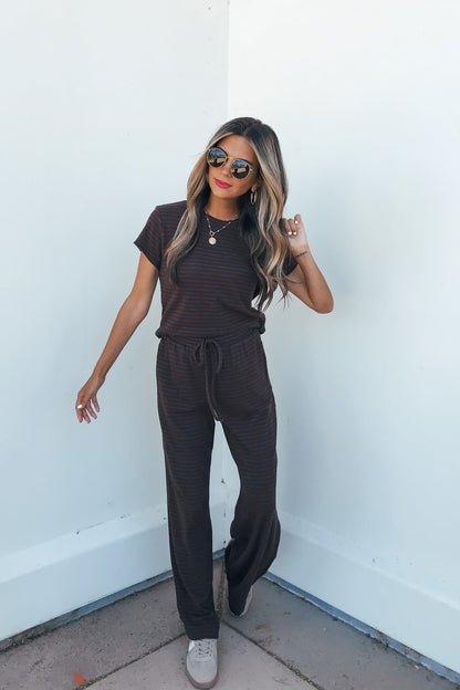 Woman stands against a white wall in Kacey Striped Wide Leg Pants - Brown, sunglasses, sneakers, and gold necklaces.