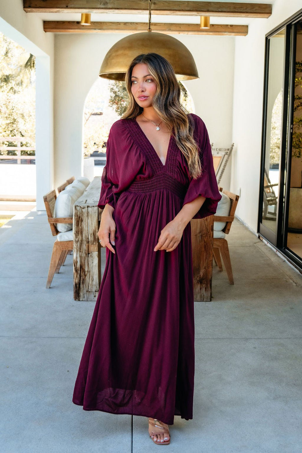 Woman wearing the Kate Burgundy Smocked Maxi Dress stands on a patio with wooden furniture and golden hanging lights.