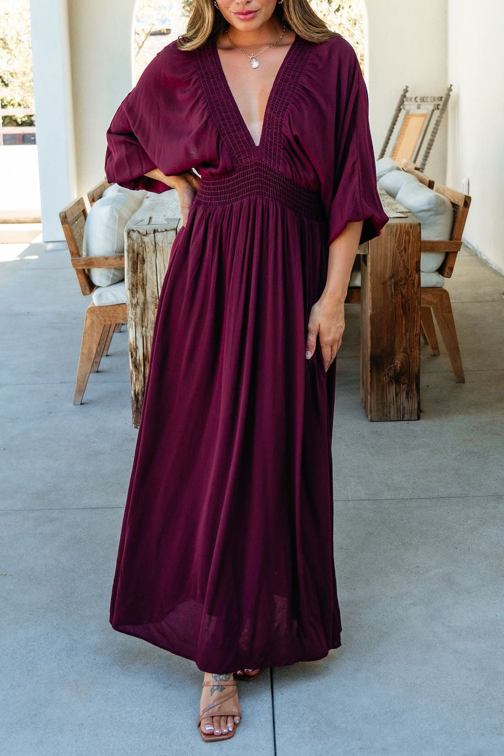 Woman in the Kate Burgundy Smocked Maxi Dress with a deep V-neck and short sleeves, standing on a patio with wooden chairs.