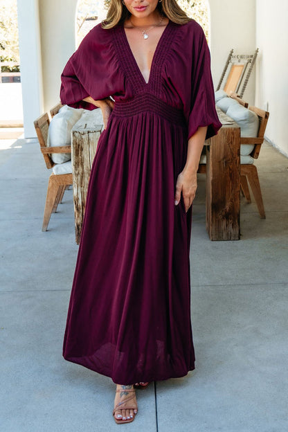 Woman in the Kate Burgundy Smocked Maxi Dress with wide sleeves, outdoors by wooden furniture. Made in USA.
