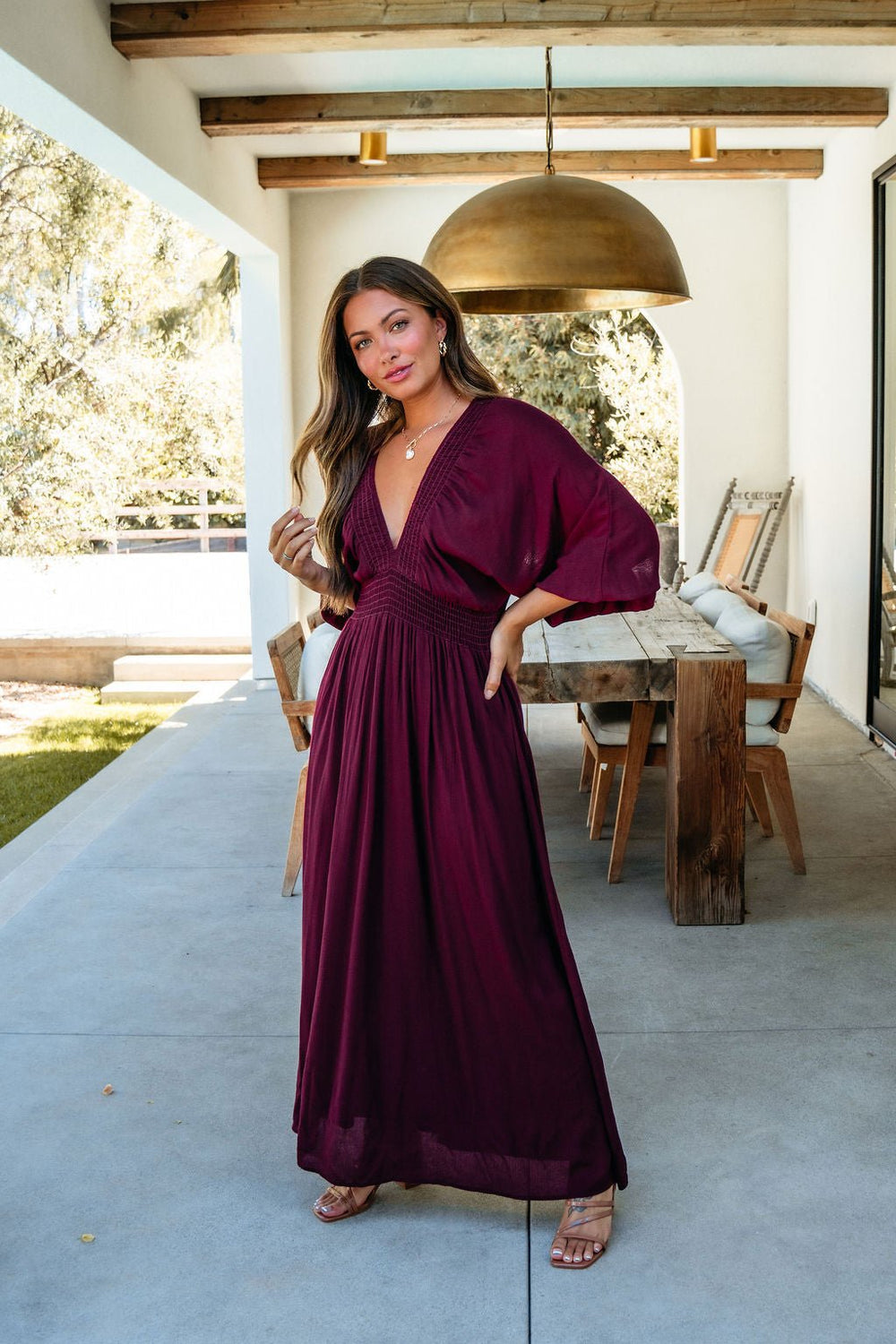 Woman in the Kate Burgundy Smocked Maxi Dress poses on a patio beside a wooden dining table under a brass light.
