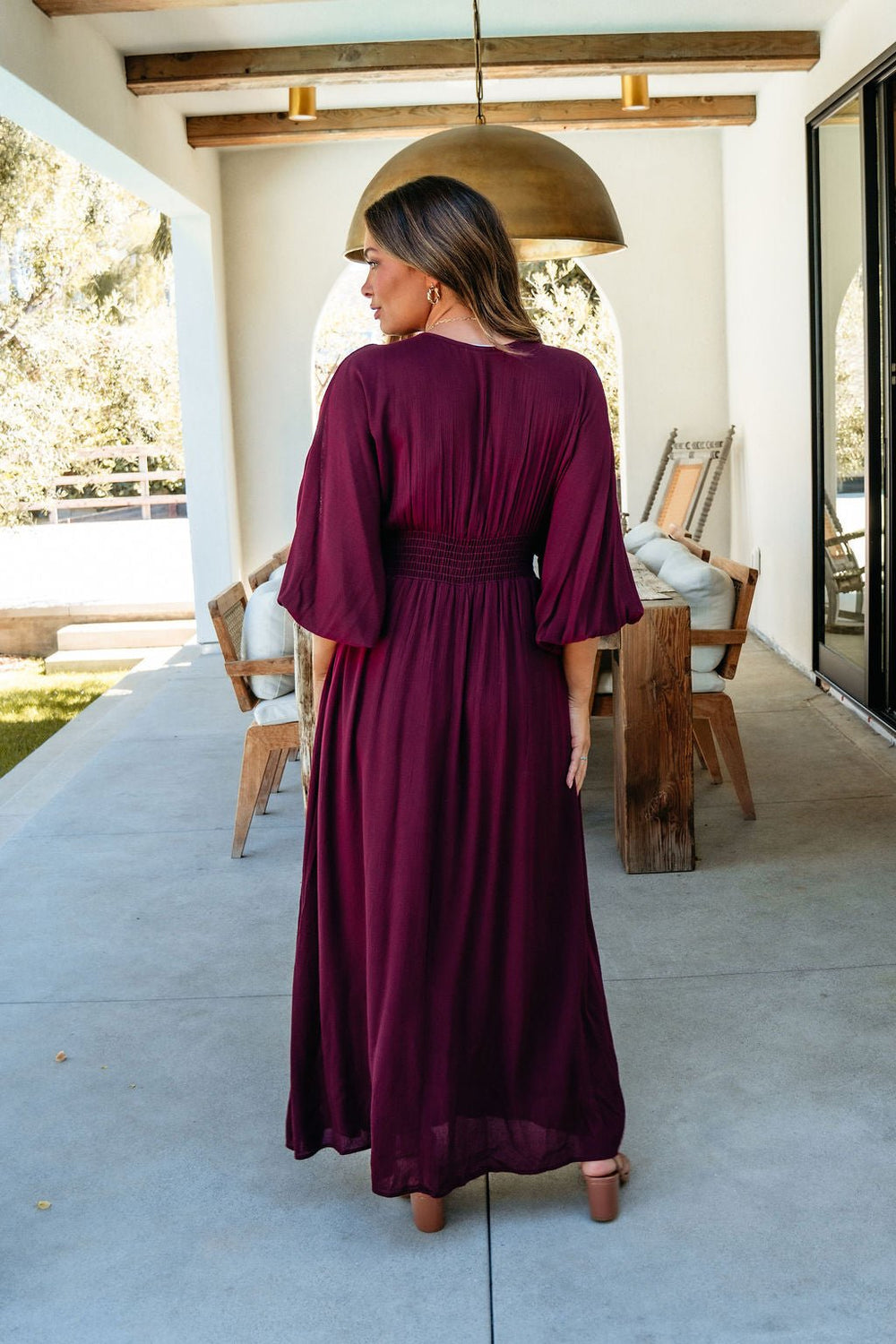A woman in the Kate Burgundy Smocked Maxi Dress stands on a patio with wooden furniture and greenery behind her.