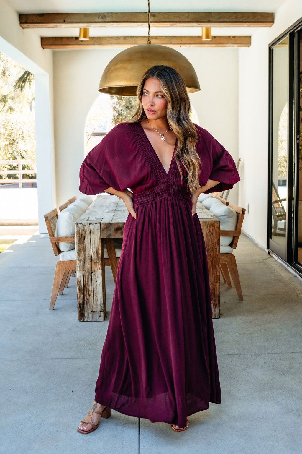 Woman in the Kate Burgundy Smocked Maxi Dress stands on a patio, looking to the side with hands on hips.
