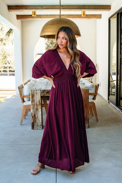 Woman in the Kate Burgundy Smocked Maxi Dress stands on a patio, looking to the side with hands on hips.