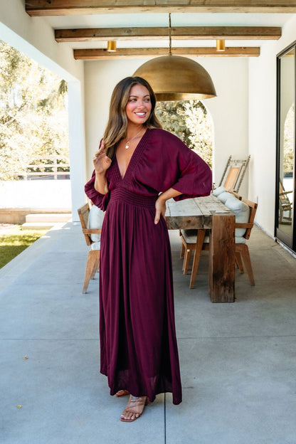 A woman smiles on a covered patio wearing the Kate Burgundy Smocked Maxi Dress, made in the USA.