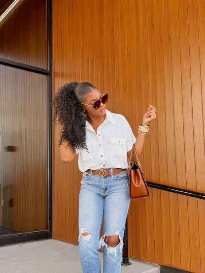 Woman in sunglasses poses by a wood-paneled wall, wearing the Keep It Simple White Button Down Shirt, ripped jeans, and a brown bag.
