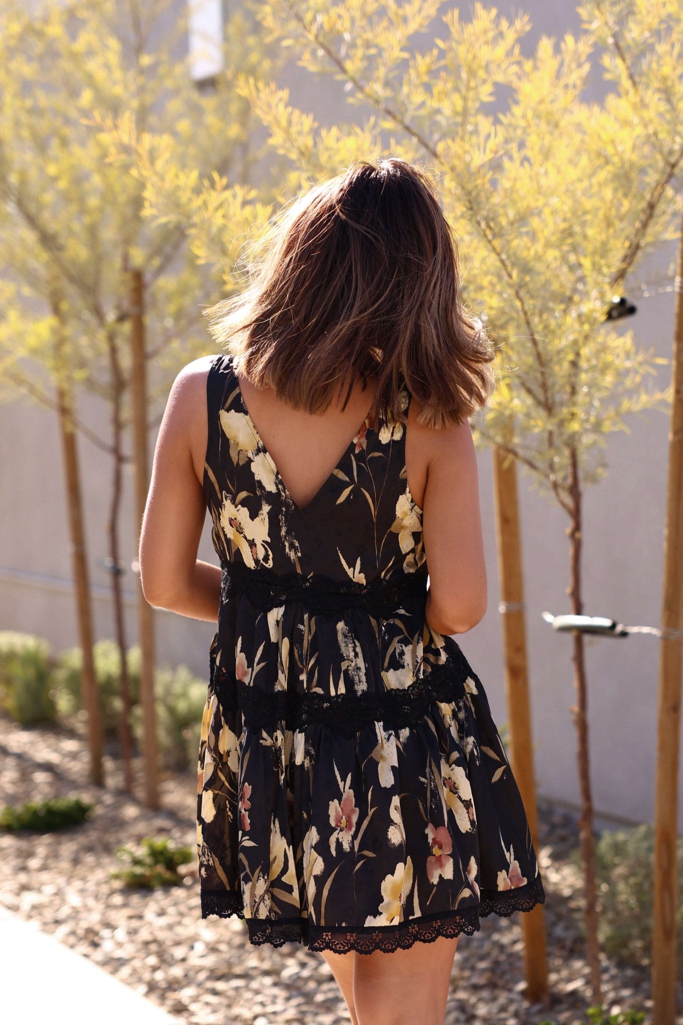 A woman in a Kenna Lace Black Print Satin Mini Dress stands outside, her back to the camera amid young trees and sunlight.