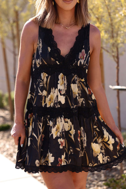 Woman in the Kenna Lace Black Print Satin Mini Dress, sleeveless with lace detail, stands outdoors in sunlight with trees behind her.