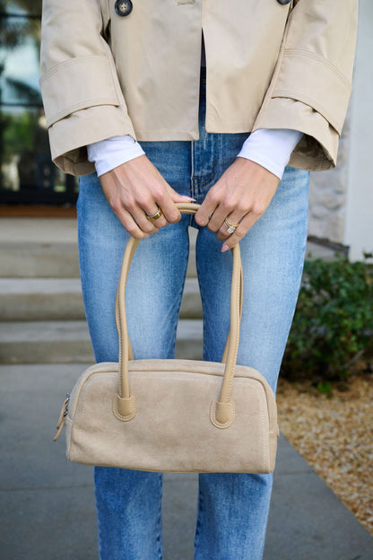 Someone in a beige coat and blue jeans holds the Khaki Faux Suede Baguette Bag with both hands in front of them.