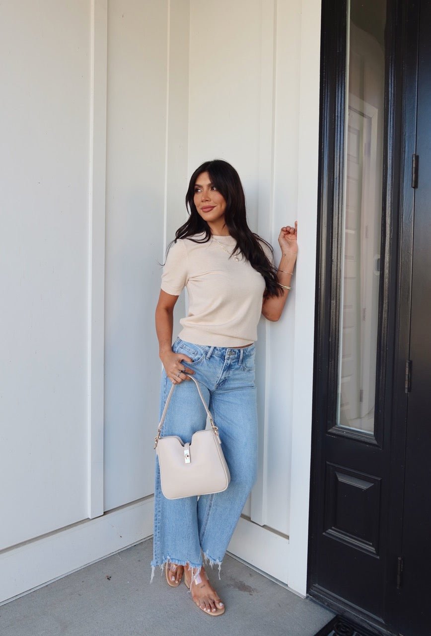 Woman in the Kiersten Champagne Short Sleeve Sweater, light blue jeans, and sandals stands by a black door with a beige purse, smiling.