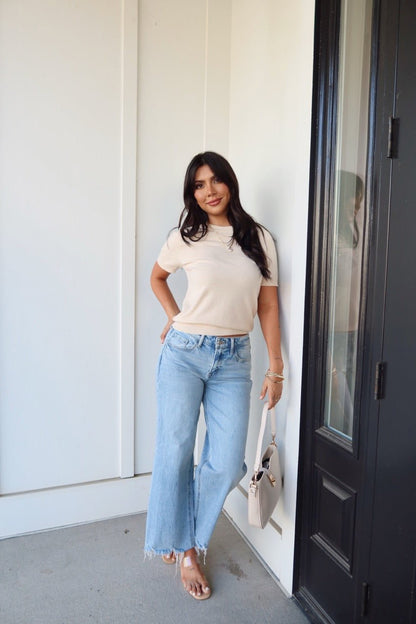 Woman with long dark hair, wearing the Kiersten Champagne Short Sleeve Sweater and blue jeans, smiles by a door holding a beige handbag.