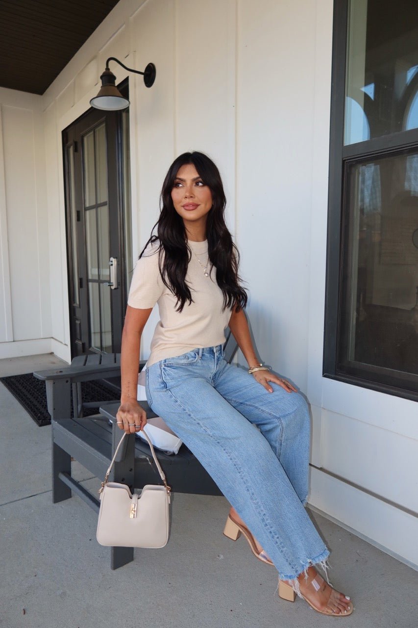 Woman with long dark hair wears the Kiersten Champagne Short Sleeve Sweater and blue jeans, sitting on a bench with a light bag.