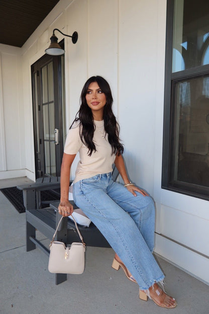 Woman with long dark hair wears the Kiersten Champagne Short Sleeve Sweater and blue jeans, sitting on a bench with a light bag.