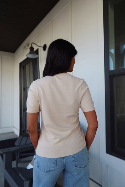 A woman with dark hair stands outside, facing away, wearing the Kiersten Champagne Short Sleeve Sweater and light blue jeans.