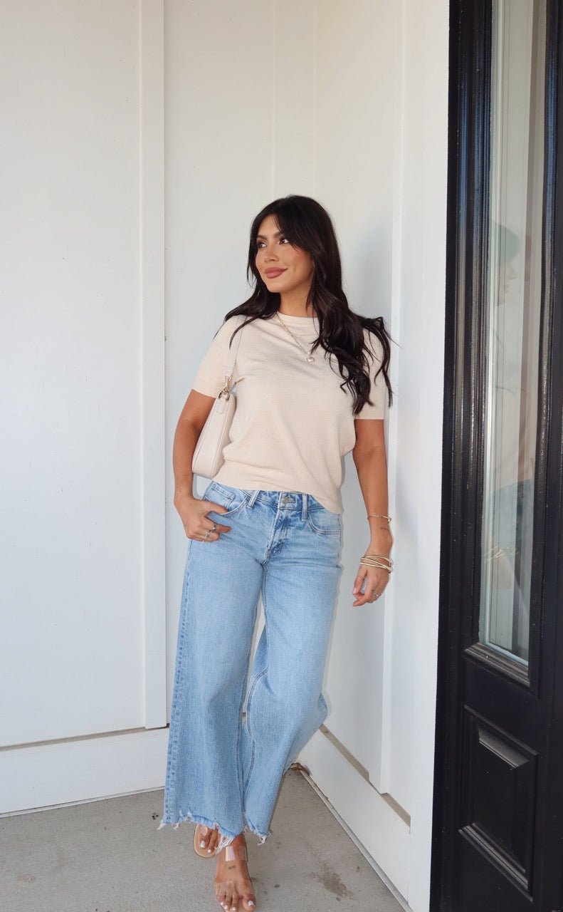 Wearing the Kiersten Champagne Short Sleeve Sweater, a woman in light blue jeans stands by a white wall, looking to the side.