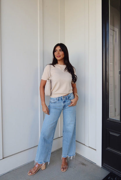 Woman in front of a white wall wears the Kiersten Champagne Short Sleeve Sweater, light blue wide-leg jeans, and clear-heeled sandals.