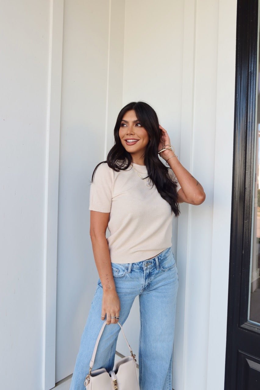 Woman with long dark hair wears the Kiersten Champagne Short Sleeve Sweater and light blue jeans, holding a beige handbag by a white wall.