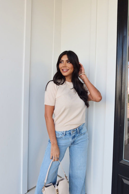 Woman with long dark hair wears the Kiersten Champagne Short Sleeve Sweater and light blue jeans, holding a beige handbag by a white wall.