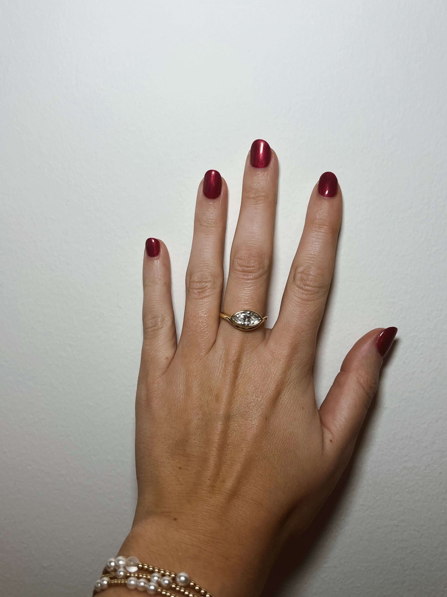 A hand with red nails wears the Kinsey Designs Griffin Gold Solitaire Ring and a pearl bracelet, set against a plain white background.