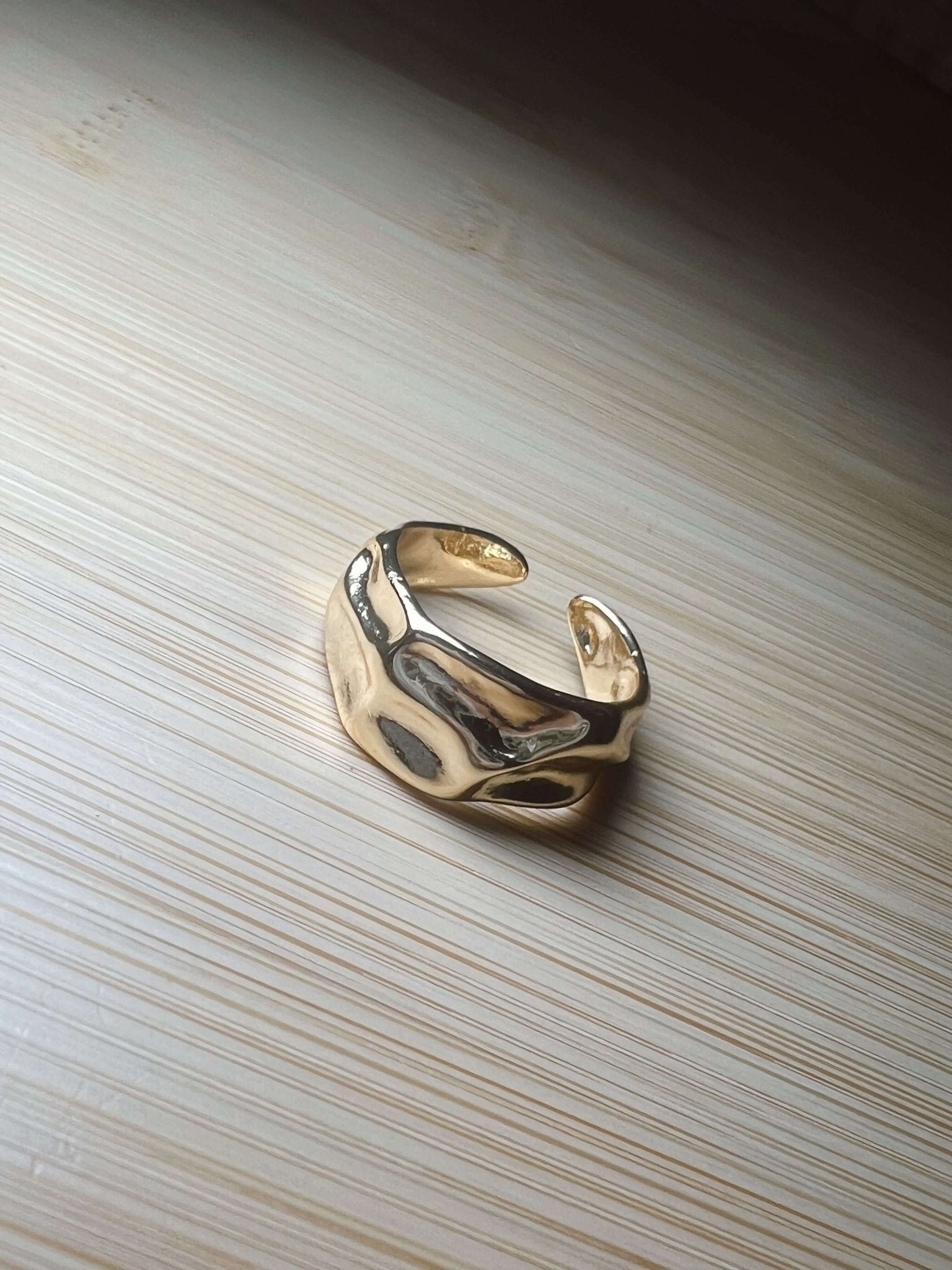 The Kinsey Designs Sybil Gold Molten Ring, a shiny 16k gold open band, rests on a light wooden surface.