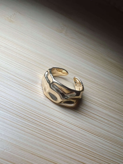 The Kinsey Designs Sybil Gold Molten Ring, a shiny 16k gold open band, rests on a light wooden surface.