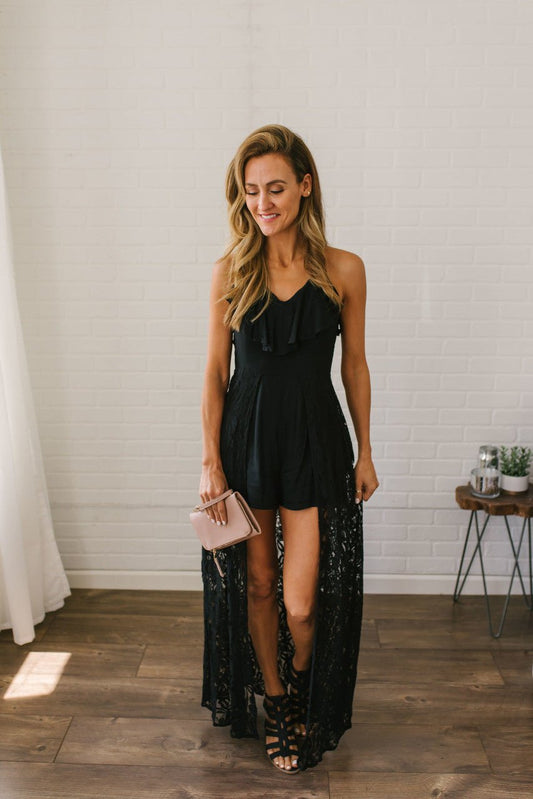 Woman wears the Kiss Me Goodnight Maxi Romper - Black, holding a blush clutch on a wooden floor by a white brick wall.