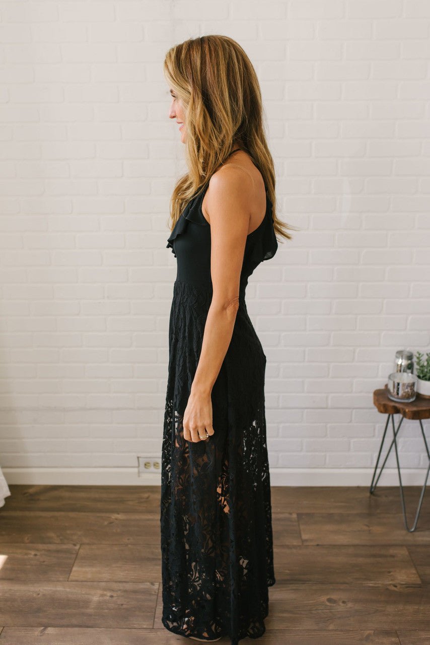 Woman in the Kiss Me Goodnight Maxi Romper - Black stands in profile in a bright room with white brick walls and wood floors.