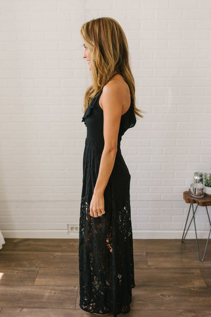 Woman in the Kiss Me Goodnight Maxi Romper - Black stands in profile in a bright room with white brick walls and wood floors.